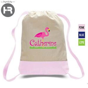 May include: A beige drawstring backpack with a pink bottom panel and matching pink shoulder straps. The front features a graphic of a pink flamingo standing on green grass above the name Catherine printed in a stylized pink script font.