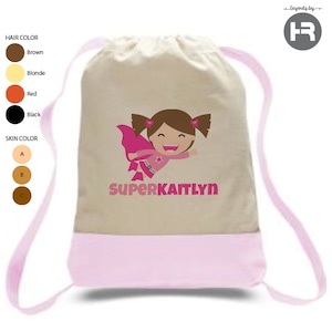 May include: A beige drawstring bag with pink straps. The bag has a pink bottom and a graphic of a girl with brown hair wearing a pink cape and a star on her shirt. The text "SUPERKAITLYN" is printed below the graphic.