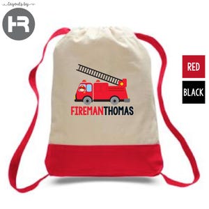 May include: A beige drawstring backpack with a red bottom panel and red shoulder straps. The front features a graphic of a red fire truck with a ladder and the text FIREMANTHOMAS printed in red and black lettering below the vehicle.