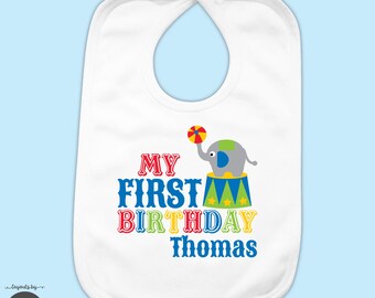 May include: A white baby bib featuring a colorful graphic of a gray elephant balancing a multi-colored ball on a circus pedestal. The text reads MY FIRST BIRTHDAY Thomas in red, blue, green, and yellow decorative lettering.