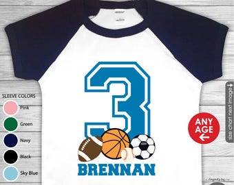 Può includere: Maglietta bianca con un numero 3 blu, una palla da baseball, una palla da basket, una palla da football americano e una palla da baseball. Il testo "BRENNAN" è sotto le palle da sport. La maglietta ha maniche blu navy. Il testo "ANY AGE" è in un cerchio rosso.