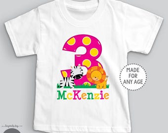 May include: A white short-sleeved t-shirt featuring a large pink number three with yellow polka dots, a cartoon zebra, and a lion. The name McKenzie is printed in green and orange letters below the animals. A circular badge reads MADE FOR ANY AGE.