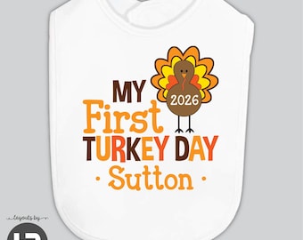 May include: A white baby bib featuring a graphic of a brown turkey with orange and yellow feathers. The turkey body displays the year 2026. The text reads MY First TURKEY DAY Sutton in brown and orange lettering with two orange dots on each side.