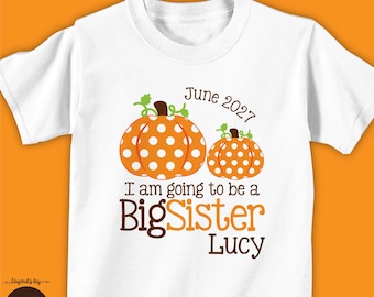 May include: White short-sleeve shirt with an orange pumpkin design. The pumpkins have white polka dots and green vines. Text on the shirt reads "I am going to be a Big Sister Lucy" and "June 2027".