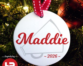 May include: A white round Christmas ornament with the name "Maddie" in red script, a megaphone graphic, and the year "2026". The ornament hangs from a red and white ribbon. Text at the bottom reads "MADE WITH ANY NAME & YEAR".