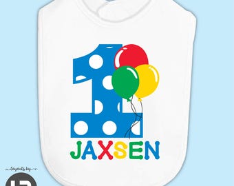 May include: A white baby bib featuring a large blue number one with white polka dots. Attached to the number are red, green, and yellow balloons with black strings. Below the number, the name JAXSEN is printed in colorful letters.