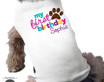 May include: A white pet shirt featuring the text "my first birthday Sophie" in teal, pink, and yellow, accompanied by a brown paw print graphic. The design is centered on the back of the garment, which is worn by a furry animal.