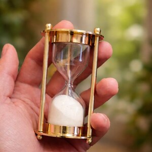 Könnte beinhalten: Eine goldfarbene Sanduhr mit klarem Glas und weißem Sand. Die Sanduhr wird in einer Hand gehalten, mit einem verschwommenen grünen und braunen Hintergrund. Die Sanduhr hat ein klassisches Design mit einer oberen und unteren Kammer, die durch einen schmalen Hals verbunden sind.