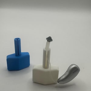 May include: An image of hearing aid accessories. A blue and white base with a white stem and a silver hearing aid with the brand name "Phonak" are visible. The accessories are designed to assist with the use and maintenance of hearing aids.