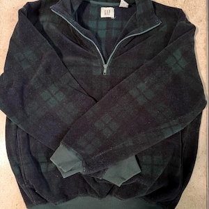 Könnte beinhalten: Ein dunkelgrüner und schwarzer Fleece-Pullover mit kariertem Muster und Viertel-Reißverschluss. Das Kleidungsstück hat einen hohen Kragen, lange Ärmel mit gerippten Bündchen und einen gerippten Saum. Ein weißes GAP-Markenetikett ist am Innenkragen sichtbar.