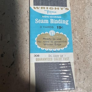 Könnte beinhalten: Eine Packung Wright's Trims Nahtband in Dunkelgrau. Die Packung ist mit "NON-SHRINK" gekennzeichnet und enthält 2,74 Meter Stoff. Der Text "GARANTIERTE FARBECHT" ist ebenfalls sichtbar.