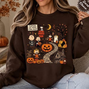 May include: A dark brown crewneck sweatshirt featuring a colorful Peanuts Halloween graphic. The design includes characters in costumes, a large jack-o'-lantern, a stone path, a house, and a sign that reads PUMPKINS, HAY-RIDES, CORN MAZE.