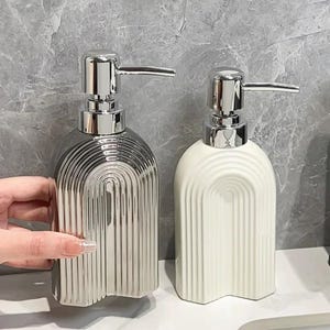 May include: Two ceramic soap dispensers with chrome pumps. One is silver with vertical grooves and an arched design. The other is white with a similar grooved pattern. Both have a modern, minimalist aesthetic.