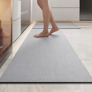 May include: A light grey kitchen mat with a textured surface and a black border. A second, smaller mat is visible in the background. The mats are placed on a tiled floor. The image shows a person standing on the mat.