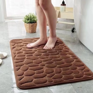 May include: A brown rectangular bath mat with a pebble design. The mat is made of a soft material and is placed on a tiled floor. A person is standing on the mat.