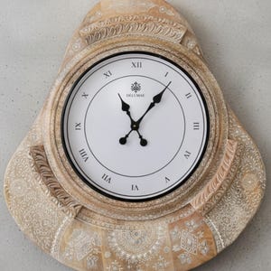 May include: A decorative wall clock with a light brown, carved wooden frame. The clock face is white with black Roman numerals and black hands. The frame features intricate floral and geometric patterns.
