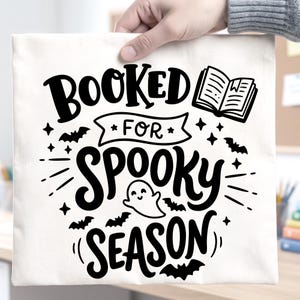 May include: A white square fabric tote bag featuring black screen-printed text that reads "BOOKED FOR SPOOKY SEASON". The design includes a small open book, a smiling ghost, several flying bats, and decorative stars scattered around the lettering.