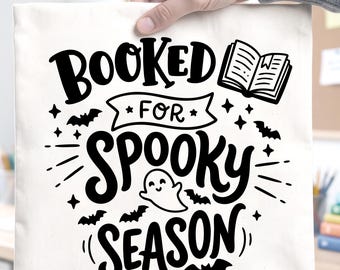 Op de afbeelding: Een witte vierkante stoffen tas met zwarte opdruk "BOOKED FOR SPOOKY SEASON". Het ontwerp bevat een klein open boek, een lachend spookje, verschillende vliegende vleermuizen en decoratieve sterren die rond de tekst zijn verspreid.