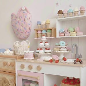 Peut inclure: Une scène de cuisine de jeu fantaisiste avec des aliments au crochet. Comprend des cornets de glace, des macarons, des beignets, des gâteaux et des fraises. La cuisine est blanche et rose, avec un panier en osier et un four rose. Une manique rose est accrochée au mur.