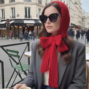 Può includere: Un set di cappuccio e sciarpa in maglia rossa con un grande dettaglio a fiocco. La sciarpa pende davanti a un blazer grigio. La persona indossa occhiali da sole neri.