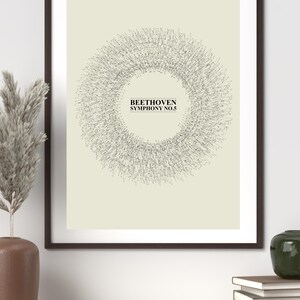 Puede incluir: Impresión artística enmarcada con un diseño circular compuesto por líneas finas, con el texto "BEETHOVEN SYMPHONY NO.5" en el centro. La obra de arte está sobre un fondo beige claro dentro de un marco marrón oscuro. La impresión se exhibe en una estantería de madera.