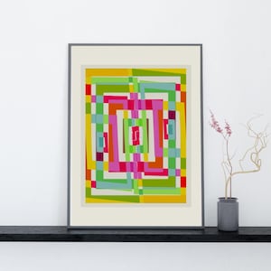 Op de afbeelding: Ingelijste abstracte kunstprint met een kleurrijk geometrisch ontwerp. Het kunstwerk toont een levendige opstelling van rechthoeken en vierkanten in groen, geel, roze en blauw. De ingelijste print wordt tentoongesteld op een zwarte plank tegen een witte muur.