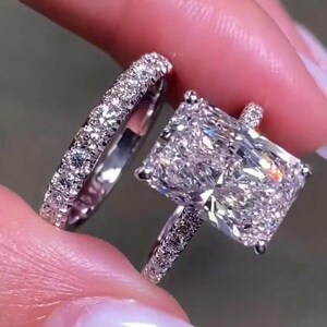 Könnte beinhalten: Ein silberfarbenes Verlobungsring-Set mit einem großen, rechteckigen Stein im Radiant-Schliff und einem passenden Ring, beide besetzt mit zahlreichen kleinen, klaren, funkelnden Steinen entlang der Ringschienen und der Hauptfassung.