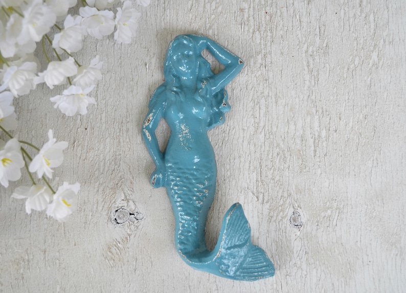 May include: A light blue mermaid wall hook with a distressed finish. The mermaid is holding her hand to her head and has a long tail.