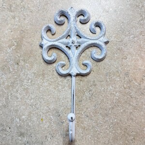 May include: A wall-mounted hook featuring a white, distressed finish with a decorative, symmetrical scrollwork design at the top. The metal piece has a central vertical stem that extends downwards into a single curved hook with a rounded tip.