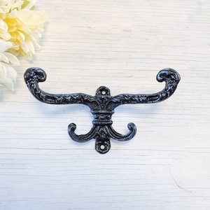 May include: Black metal coat hook with a decorative scroll design. The hook has two prongs and is mounted on a white wall.