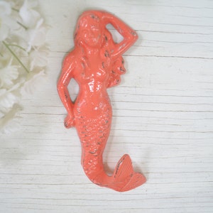May include: Coral pink mermaid wall hook with a textured finish. The mermaid is holding her hand to her head and has a long flowing tail.
