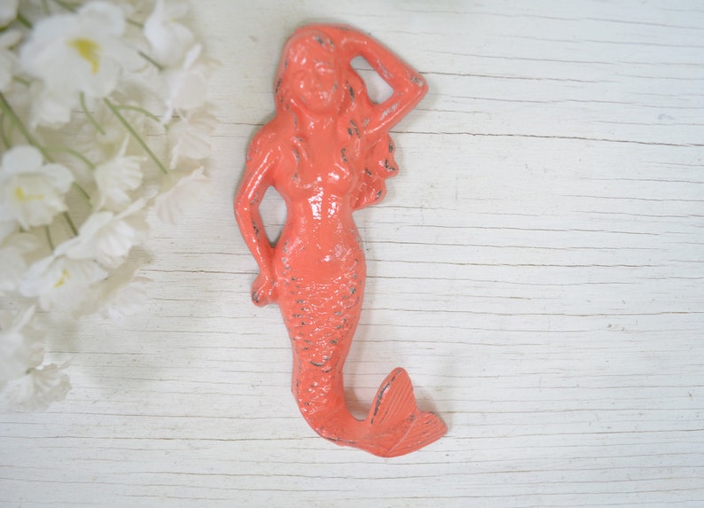 May include: Coral pink mermaid wall hook with a textured finish. The mermaid is holding her hand to her head and has a long flowing tail.