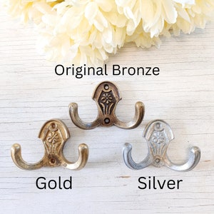 May include: Three double-hook wall hangers in different finishes: original bronze, gold, and silver. Each hook features a decorative floral design.