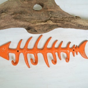 May include: Orange metal fish skeleton wall hook with five hooks. The hook is shaped like a fish skeleton with a tail and a head. The hook is mounted on a white background.