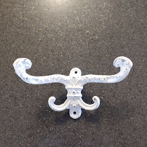 May include: White, distressed-finish metal wall hook with a fleur-de-lis design. The hook has two curved arms and a lower decorative element with two additional hooks. The hardware is designed for wall mounting.