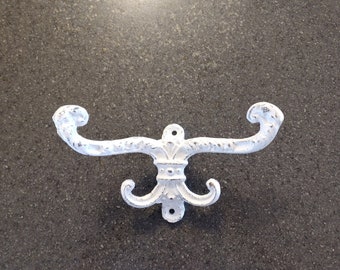 May include: White, distressed-finish metal wall hook with a fleur-de-lis design. The hook has two curved arms and a lower decorative element with two additional hooks. The hardware is designed for wall mounting.