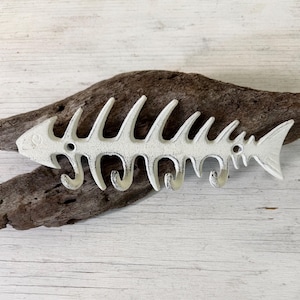 May include: A white, distressed-finish cast iron wall hook shaped like a fish skeleton. The design features a long, horizontal spine with multiple upward-curving ribs and four integrated hooks along the bottom edge for hanging items.