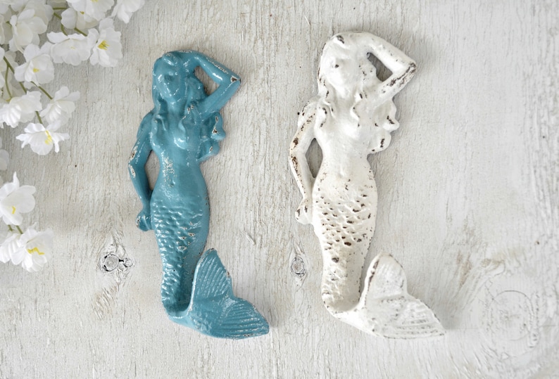 May include: Two mermaid-shaped hooks, one in teal blue and one in white, with distressed paint finishes. They are mounted on a white wooden surface.