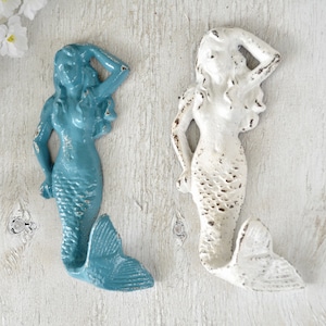 May include: Two mermaid-shaped hooks, one in teal blue and one in white, with distressed paint finishes. The hooks are mounted on a white wooden surface.