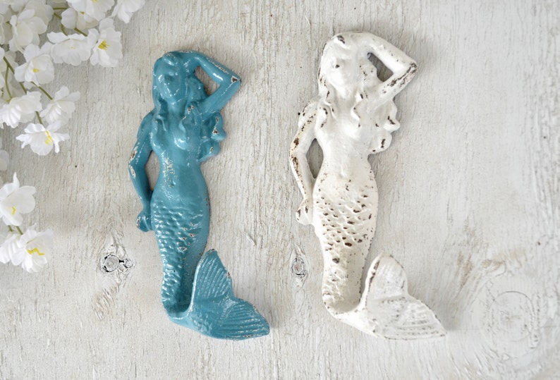 May include: Two mermaid-shaped hooks, one in teal blue and one in white, with distressed paint finishes. The hooks are mounted on a white wooden surface.