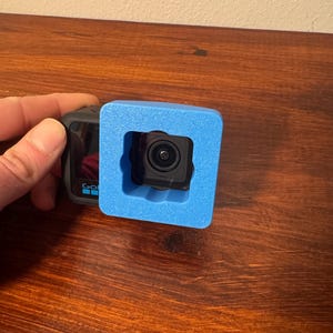 May include: A black action camera with a lens is held within a bright blue foam protective case. The camera is positioned on a dark wooden surface. The camera has the word "GO" printed on the side.