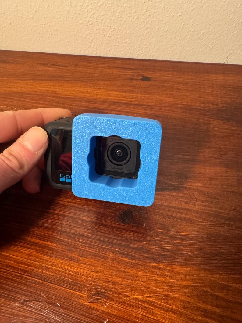 May include: A black action camera with a lens is held within a bright blue foam protective case. The camera is positioned on a dark wooden surface. The camera has the word "GO" printed on the side.
