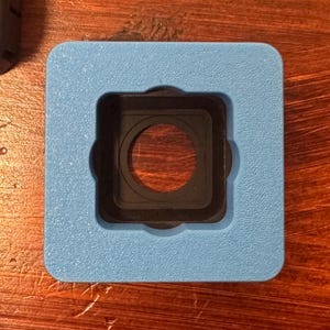 May include: A square, light blue object with a black, circular center. The object has a square cutout in the center, with a circular opening. The object is likely a camera accessory or part.
