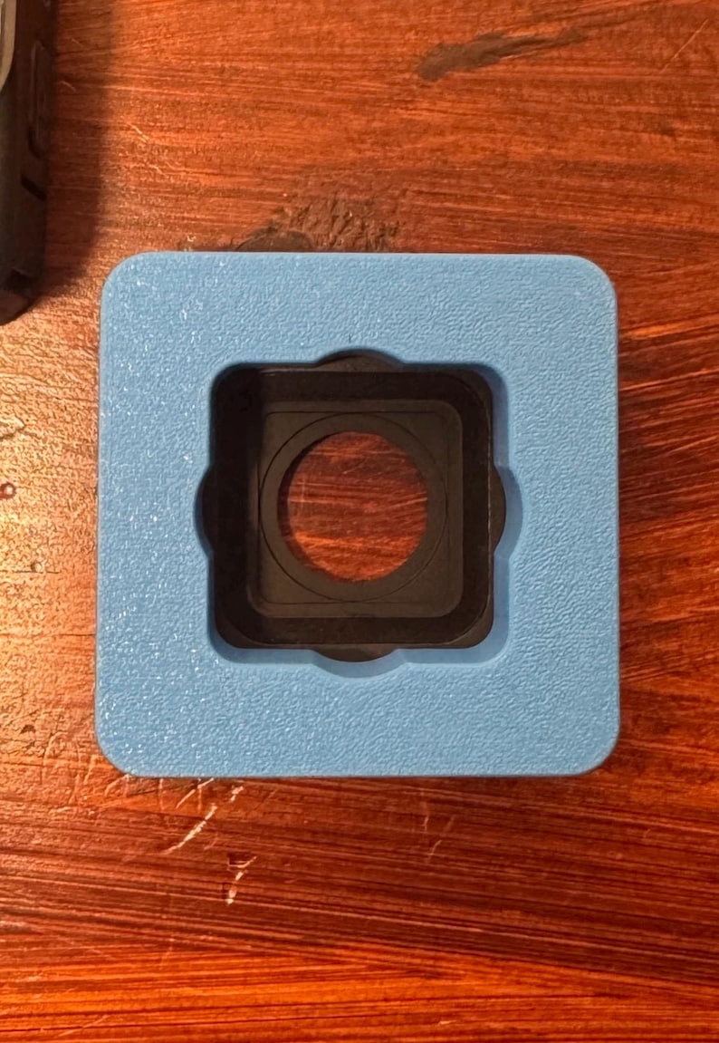 May include: A square, light blue object with a black, circular center. The object has a square cutout in the center, with a circular opening. The object is likely a camera accessory or part.