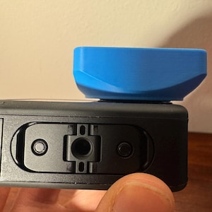 May include: A black action camera with a bright blue accessory attached. The accessory is rectangular with curved edges. The camera has a series of ports and buttons.