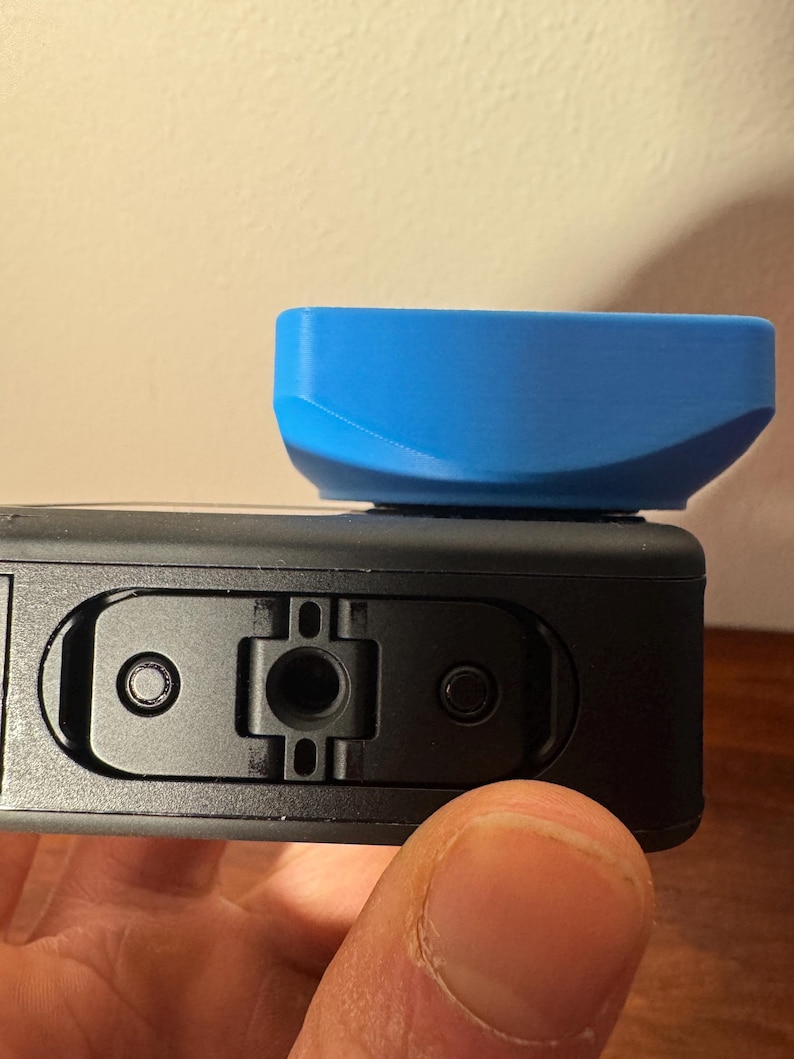 May include: A black action camera with a bright blue accessory attached. The accessory is rectangular with curved edges. The camera has a series of ports and buttons.
