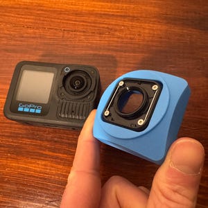 May include: A black GoPro camera with a screen and lens, next to a light blue camera housing. The camera has the GoPro logo on the front. The housing has a circular opening for the lens.