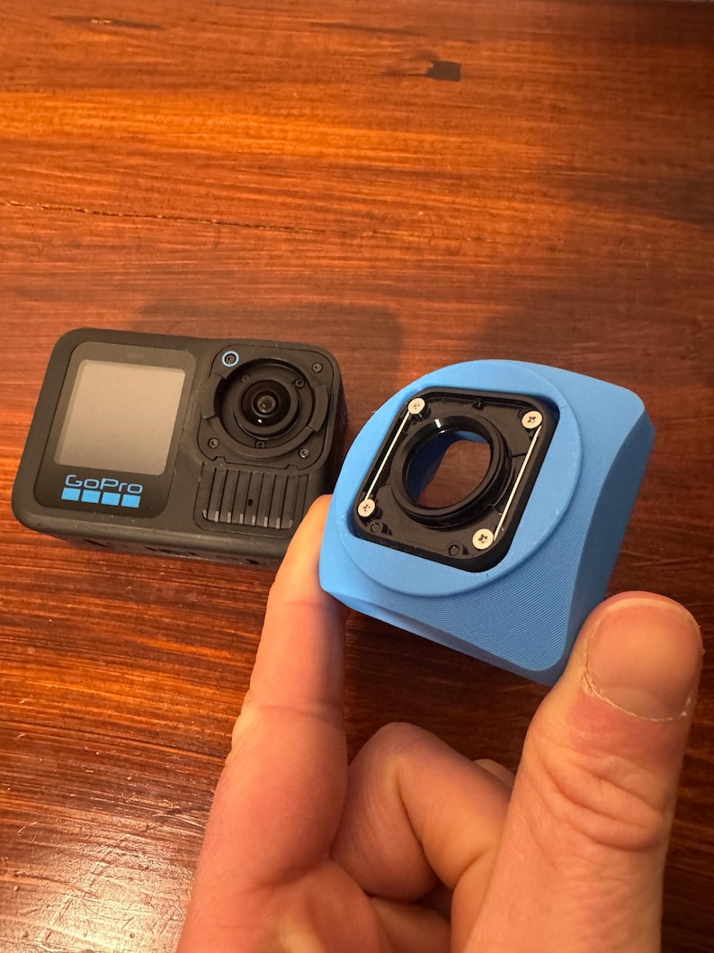 May include: A black GoPro camera with a screen and lens, next to a light blue camera housing. The camera has the GoPro logo on the front. The housing has a circular opening for the lens.