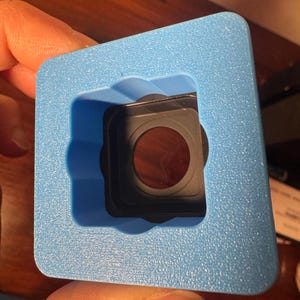 May include: A light blue foam insert with a square cutout, holding a black and brown object. The object has a circular lens. The foam has a textured surface.