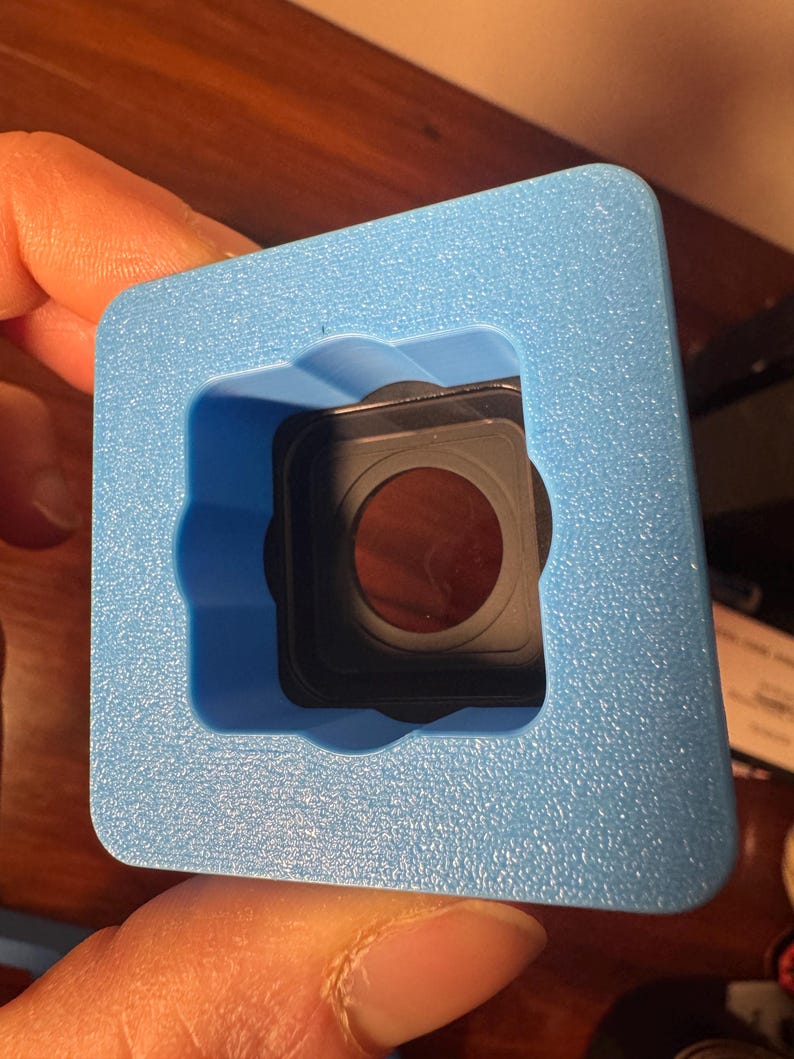 May include: A light blue foam insert with a square cutout, holding a black and brown object. The object has a circular lens. The foam has a textured surface.